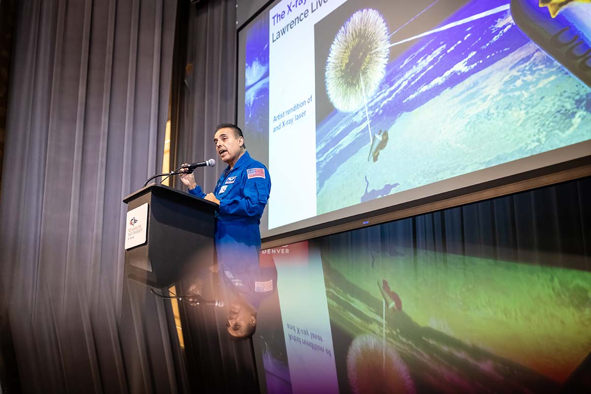 Astronaut José Hernández, wearing a blue NASA flight suit, speaks at a podium during a presentation at Metropolitan State University of Denver. A large screen behind him displays a scientific diagram, possibly an artist's rendition of an X-ray laser."